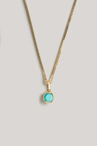 A gold chain necklace with a small round bezel-set turquoise pendant, displayed against a neutral light gray background.