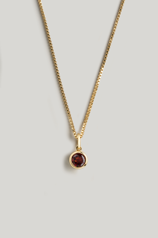 A gold chain necklace with a small round bezel-set dark red gemstone pendant, displayed against a neutral light gray background.