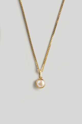 A gold chain necklace with a small round bezel-set pearl pendant, displayed against a neutral light gray background.