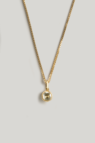 A gold chain necklace with a small round bezel-set pale yellow-green gemstone pendant, displayed against a neutral light gray background.