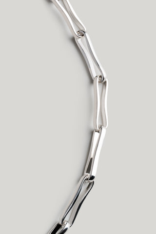 A close-up of a polished silver chain with elongated, geometric links arranged in a curved line against a solid light gray background.