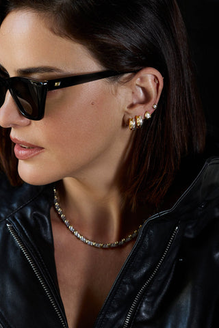 Model in sunglasses and a leather jacket wearing stacked Romy huggies, a pear-shaped stud and a two-tone tennis necklace