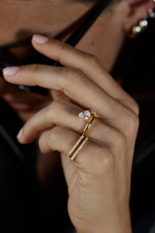 Gold plated Livia ring stacked with a second pave band beneath a pear-shaped clear stone ring, hand raised to sunglasses