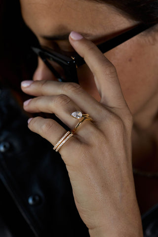 A close-up of a person's hand wearing three gold rings, including a diamond engagement ring and two stacked band rings, while holding black-framed glasses.
