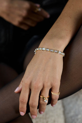 A close-up of a person's hand wearing a delicate diamond bracelet with gold-toned settings, along with gold rings, set against dark tights and a soft neutral background.