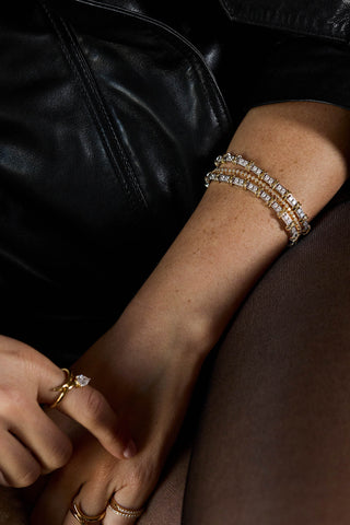 A close-up of a person's wrist and hand wearing multiple delicate gold bracelets adorned with clear stones, paired with a black leather jacket and sheer brown tights.