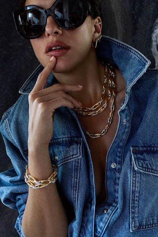 Model in sunglasses and a denim shirt, finger to her lip, wearing layered two-tone Allegra necklaces and bracelets