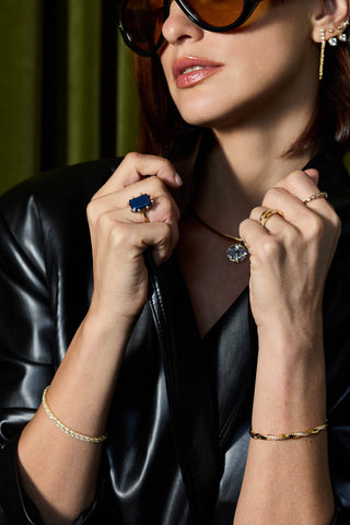 A close-up lifestyle shot of a woman wearing multiple gold jewelry pieces, including a gold chain necklace with a clear stone pendant, a gold bracelet, a gold ring with a deep blue rectangular stone, and a gold ear cuff, while wearing a black leather jacket.
