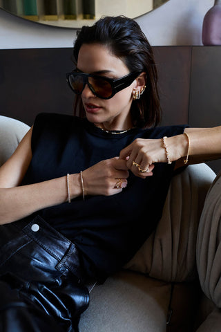 Model in sunglasses and a black top on a sofa wearing gold-tone tennis bracelets on both wrists, rings, hoops and a necklace