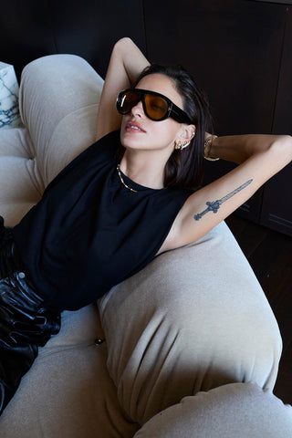 Model reclining on a beige sofa in sunglasses and a black top, wearing a gold-tone station necklace and stacked hoops