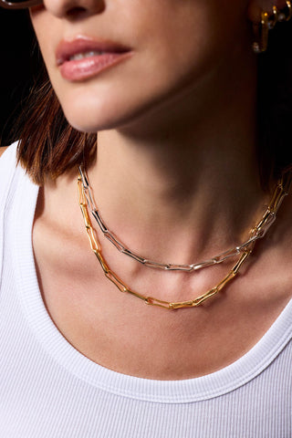 A close-up of a model wearing two layered chain necklaces, one gold and one silver, styled with a white ribbed tank top.