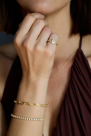 Gold plated Protective Eye ring worn with a paperclip bracelet and a heart-stone tennis bracelet, hand raised to the chin