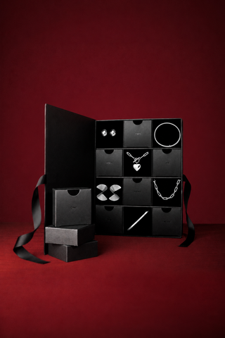 An open black jewelry gift box with multiple compartments, displayed on a dark red background. The box contains silver jewelry pieces including a heart pendant necklace, a chain necklace, earrings, and rings.