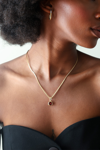 A close-up lifestyle shot of a model wearing a delicate gold chain necklace with a small round pendant featuring a dark red gemstone.