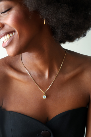 A close-up lifestyle shot of a smiling woman wearing a delicate gold chain necklace with a small round light blue pendant, paired with a black strapless top.